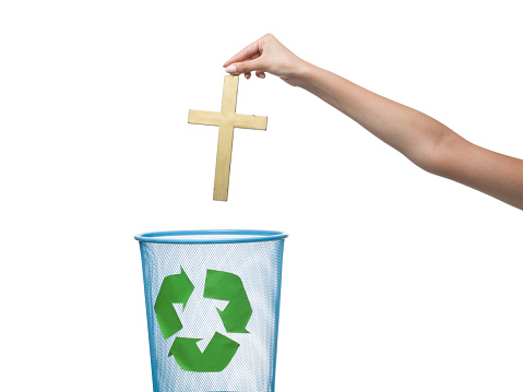 Human hand holding a gold color christianity cross over garbage bin.The bin is full of books and has a recycling sign on it.The bin is on the down left of frame while female human hand is on the right side of frame.The image was shot in studio on white background.