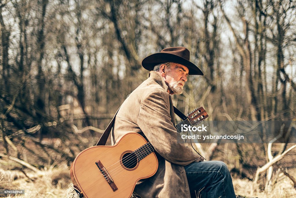 Lonely sad senior guitarist Lonely sad senior guitarist sitting outdoors in woodland with his guitar under his arm staring off into the distance with a thoughtful expression Cowboy Hat Stock Photo
