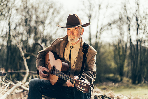 Senior man in a stylish western outfit sitting alone outdoors playing country music on an acoustic guitar