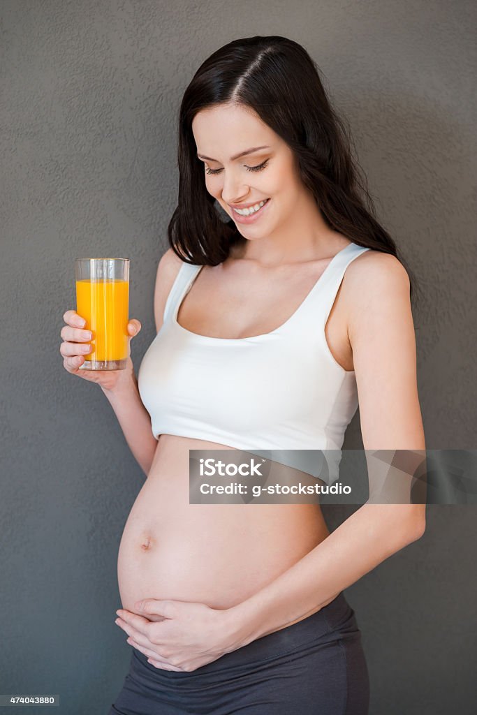 Fresh juice for my baby. Beautiful pregnant woman holding a glass of orange juice and smiling while standing against grey background 2015 Stock Photo