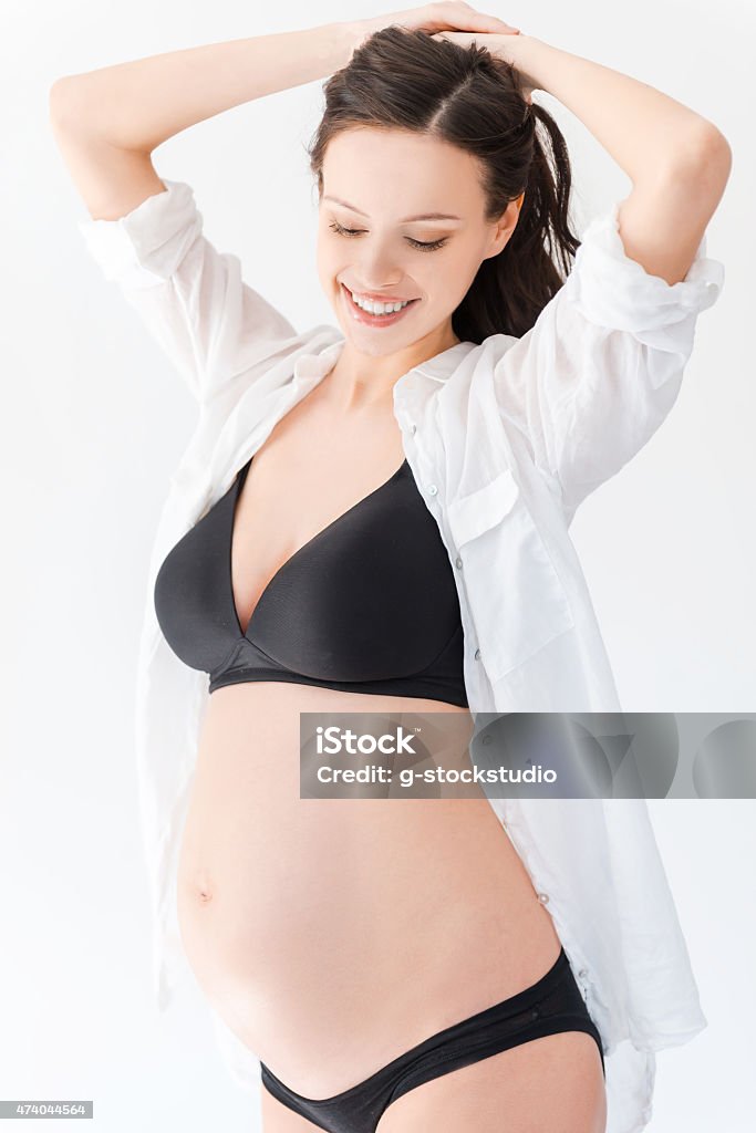 The happiest time for me. Beautiful pregnant woman keeping arms raised and smiling while standing against white background 2015 Stock Photo