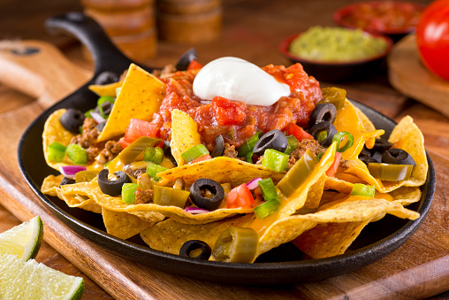 A plate of delicious tortilla nachos with melted cheese sauce, ground beef, jalapeno peppers, red onion, green onions, tomato, black olives, salsa, and sour cream with guacamole dip.