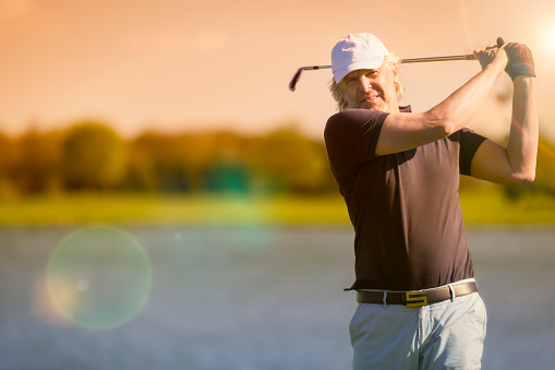 Male senior golf player swinging golf club with lake in background at sunset, with sun flare.