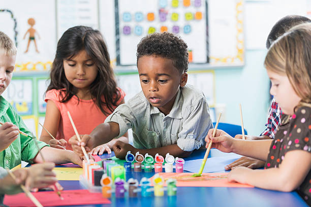 Group of preschoolers in art class painting pictures A group of preschool or kindergarten children in art class painting pictures at a table.  A little African American boy, 6 years old, is in the middle, reaching over to dip his brush in the paint. child paintbrush stock pictures, royalty-free photos & images