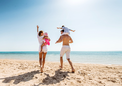 Cheerful parents enjoying at the beach with their little children. Man is carrying little boy on his shoulders. Copy space.