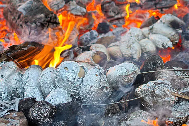 Baked potatoes covered with aluminum foil roasting in a bonfire with orange flames and firewood