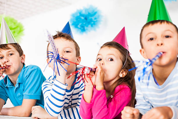 Three boys dressed up in colorful party clothing are celebrating a birthday. One child is wearing a colorful clown wig, a red nose, and a giant bow tie, the other two boys have party hats and funny sunglasses on, one of them is blowing a party blower. This is a tinted, retro style image of the childrens party.