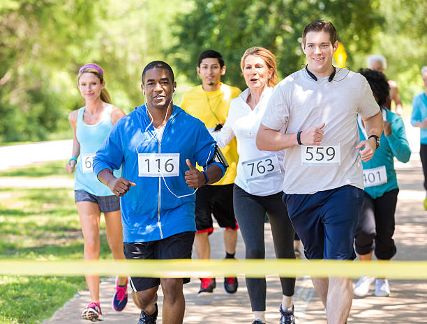 Diverse runners approaching finish line during marathon in park African American mid adult man and Caucasian mid adult man are smiling as they approach yellow finish line tape during marathon or 5k race. Runners are followed by diverse race contestants who are all trying to win race in park on sunny day. Everyone is wearing athletic clothing and a contestant number on their shirt. walking for a cause stock pictures, royalty-free photos & images