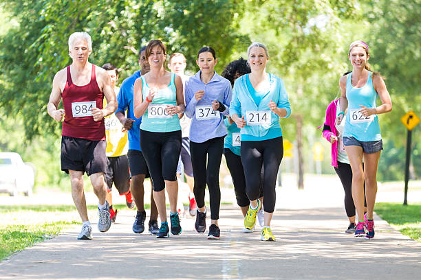 Diverse running team approaching finish line during marathon Diverse group of adults running together during marathon race in park on sunny day. They are smiling as they approach finish line. Group of runners is wearing multicolored athletic clothing and race contestant numbers. walking for a cause stock pictures, royalty-free photos & images