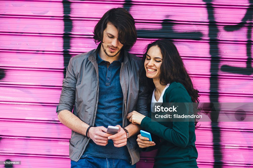 Young couple chatting on the phone Young couple having fun during they are chatting. They both looking at they phones. They standing in front of pink painted wall and smiling Couple - Relationship Stock Photo