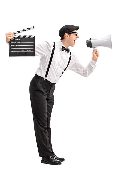 Full length profile shot of a young movie director holding a clapperboard and shouting on a megaphone isolated on white background