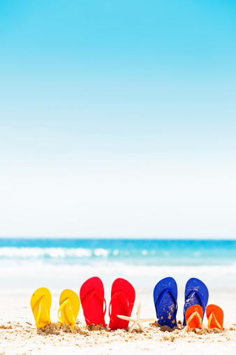 Let the family vacation fun begin! Pink, blue, yellow, and orange flipflop rubber sandals sit on a sandy beach with calm sea in the background. Plenty of copy space on the sand and sea.