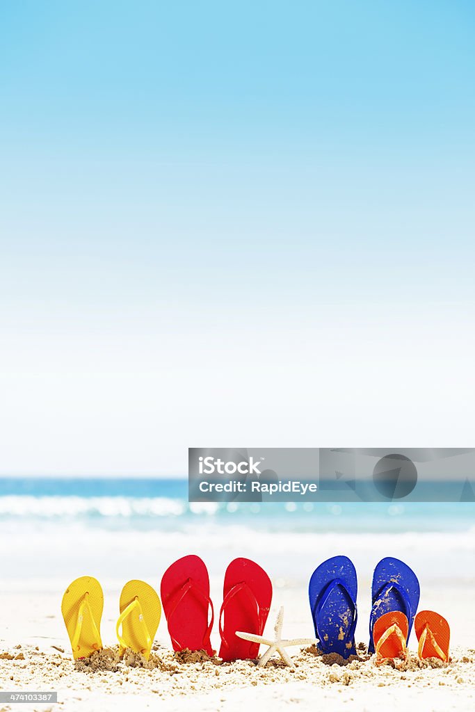 Family vacation time: four pairs of flipflops on summer beach Let the family vacation fun begin! Pink, blue, yellow, and orange flipflop rubber sandals sit on a sandy beach with calm sea in the background. Plenty of copy space on the sand and sea. Family Stock Photo