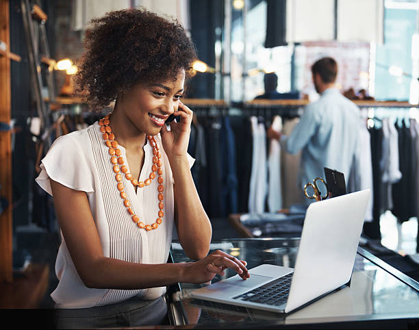 She enjoys her job A woman sitting at the desk of her clothing boutique and talking on her phone while looking at her laptop clothing store stock pictures, royalty-free photos & images