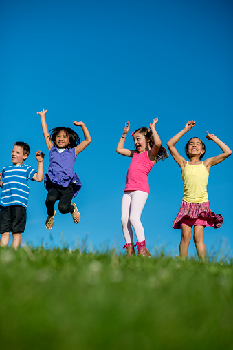 An action shot of a multi-ethnic group of elementary age children jumping in the air, outside at the park atop a grassy hill, with the blue sky in the background.