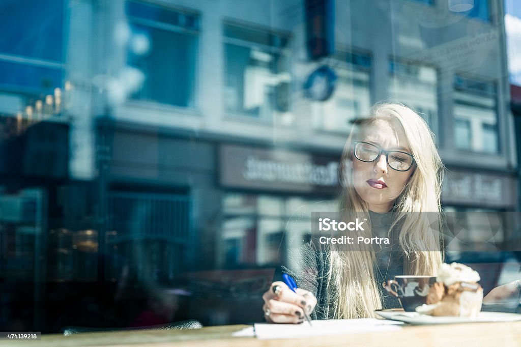 Young woman with glasses writing on blank paper having coffee Young caucasian white woman with glasses, red lipstick and long blonde hair looking down with serious face while writing with blue pen on blank white paper working on new project and having coffee and cake seen through window with sunny cityscape reflection in the horizontal glass window. Vintage looking warm and cold light photo. 2015 Stock Photo