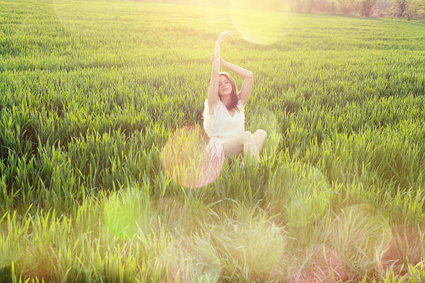 Young woman sitting in a wheat field relaxing and enjoying the light and nature with raised arms. A lot of lens flares and defocused lights flowing along her. 