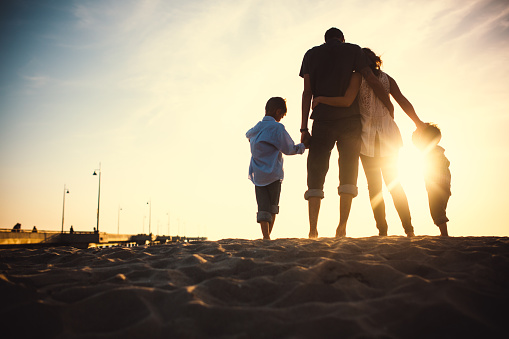 A silhouette of a beautiful young family on vacation at Venice beach, California watching the sun set over the ocean, the Venice Fishing Pier visible behind them.  The parents hug, holding the hands of their children.  Horizontal with copy space.