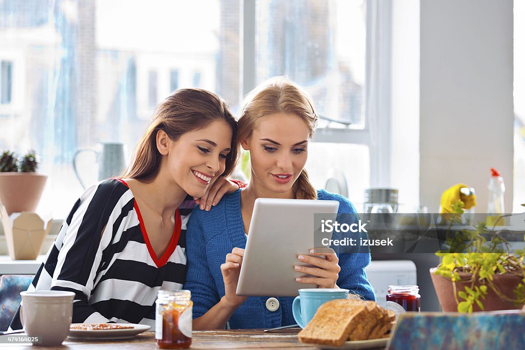 Girls using digital tablet Two cheerful friends sitting at the kitchen table and using a digital tablet together.  20-24 Years Stock Photo