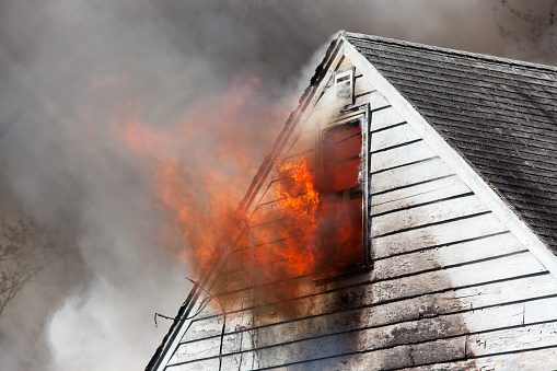 A fully engulfed old house fire with flames coming through an attic window and smoke filling the frame. Streaks in the smoke are from sunlight.