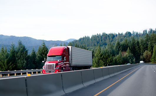 Red powerful modern professional semi-truck to haul a refrigerated trailer on the interstate highway with a concrete separation barrier against the backdrop of evergreen trees on the hills.