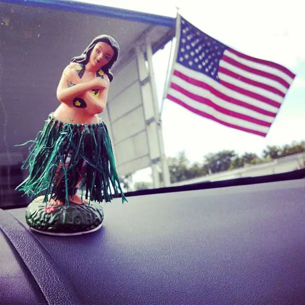 This is a square, mobilestock photograph of a vintage hula girl bobble head on a car's dashboard. In the background is the American Flag.