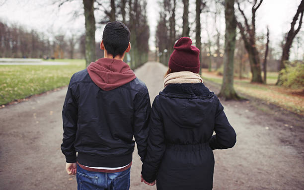 Teenage couple walking in park holding hands Rear view of teenage couple walking in park holding hands. Young man and woman in warm clothes outdoors. teen romance stock pictures, royalty-free photos & images