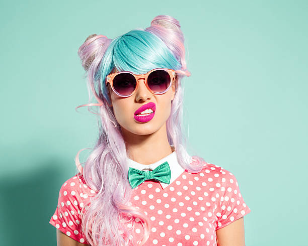 Pink hair manga style girl grimacing Portrait of manga style blue-pink hair girl wearing sunglasses and pink polka dot dress with collar and bow tie. Standing against turquoise background, looking at camera. Studio shot, one person. uncertainty photos stock pictures, royalty-free photos & images