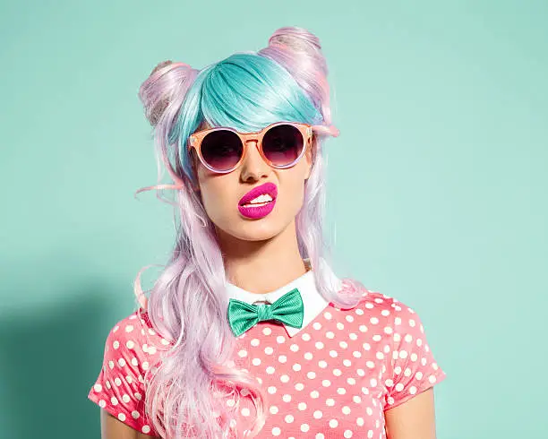 Portrait of manga style blue-pink hair girl wearing sunglasses and pink polka dot dress with collar and bow tie. Standing against turquoise background, looking at camera. Studio shot, one person.