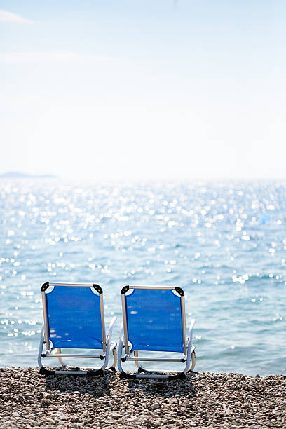 Two beach chairs in front of defocused shiny waves and bright sunlight in a Greek pebble beach.