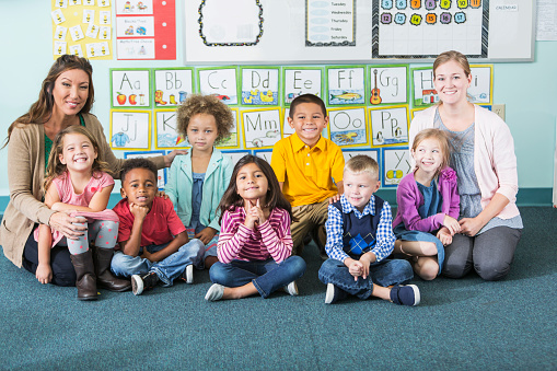 Two preschool or kindergarten teachers with a group of multiracial children in class.  They are sitting on the floor, legs crossed, smiling at the camera.  On the wall behind them is artwork with the alphabet.