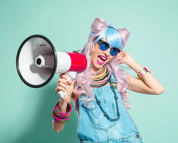 Portrait of happy manga style blue-pink hair young woman wearing denim coveralls and sunglasses, shouting into megaphone. Standing against turquoise background. Studio shot, one person.