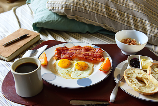 Classic breakfast, bacon, eggs, oatmeal, coffee, bagel with butter and jelly on wood serving tray set in unmade bed with leather journal and pen to one side