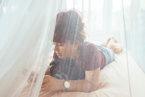 This is a horizontal, color, royalty free stock photograph of a traveling Canadian woman in her 20s. She lays across a bed covered with mosquito netting in Thailand. She looks at her phone, checking emails, texting and looking at social media. Her hair is dyed red. Photographed with a Nikon D800 DSLR camera.