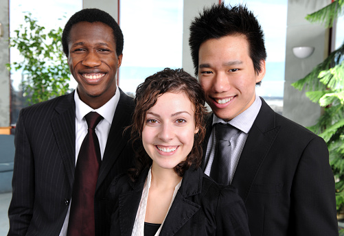 A multi-ethnic group of interns standing together in an office building - smiling and looking at the camera.