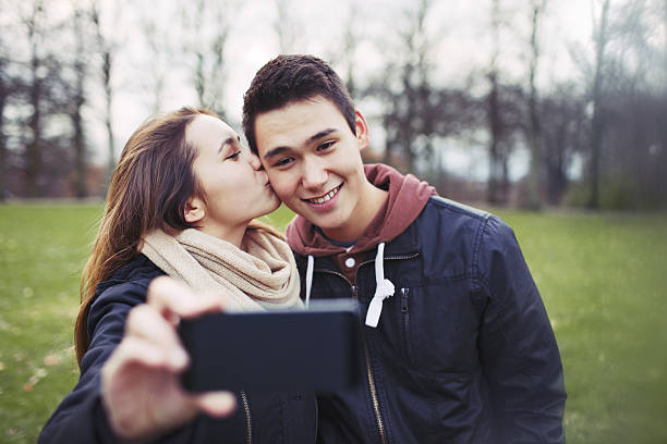 Loving teenage couple taking self portrait with a mobile phone Pretty young girl kissing her boyfriend on cheeks while taking self portrait with a mobile phone. Mixed race couple in park. cheek cell stock pictures, royalty-free photos & images