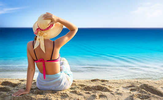 Beach vacation, young woman in sunhat and bikini sitting with her arm raised to her head enjoying looking view of beach ocean on hot summer day.