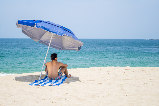 A latin young male with athletic body sitting on a towel with blue and white stripes under  a umbrella at the beach with the ocean on the background. He is facing to the ocean.