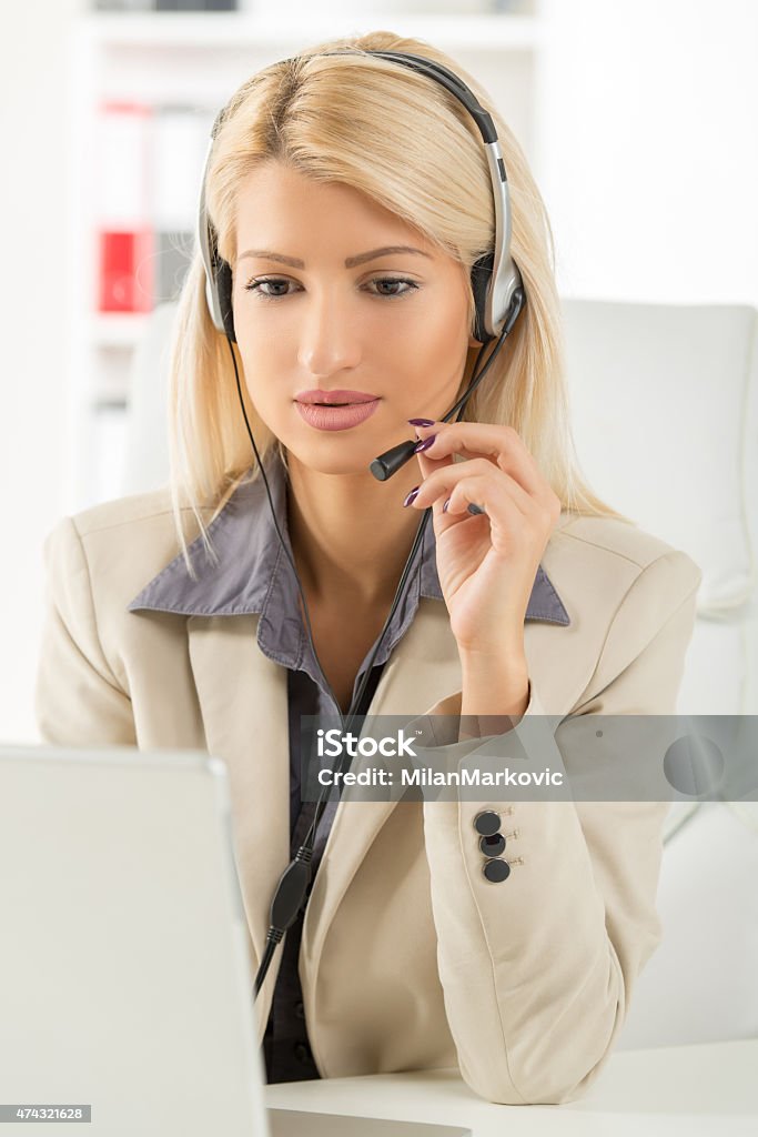 Young Call Operator Young beautiful blonde girl, elegantly dressed, sitting at an office desk in front of laptop with a headset on her head. 2015 Stock Photo