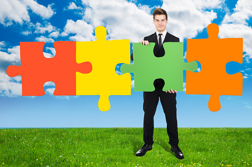 Full length portrait of confident young businessman solving jigsaw puzzle on field against cloudy sky