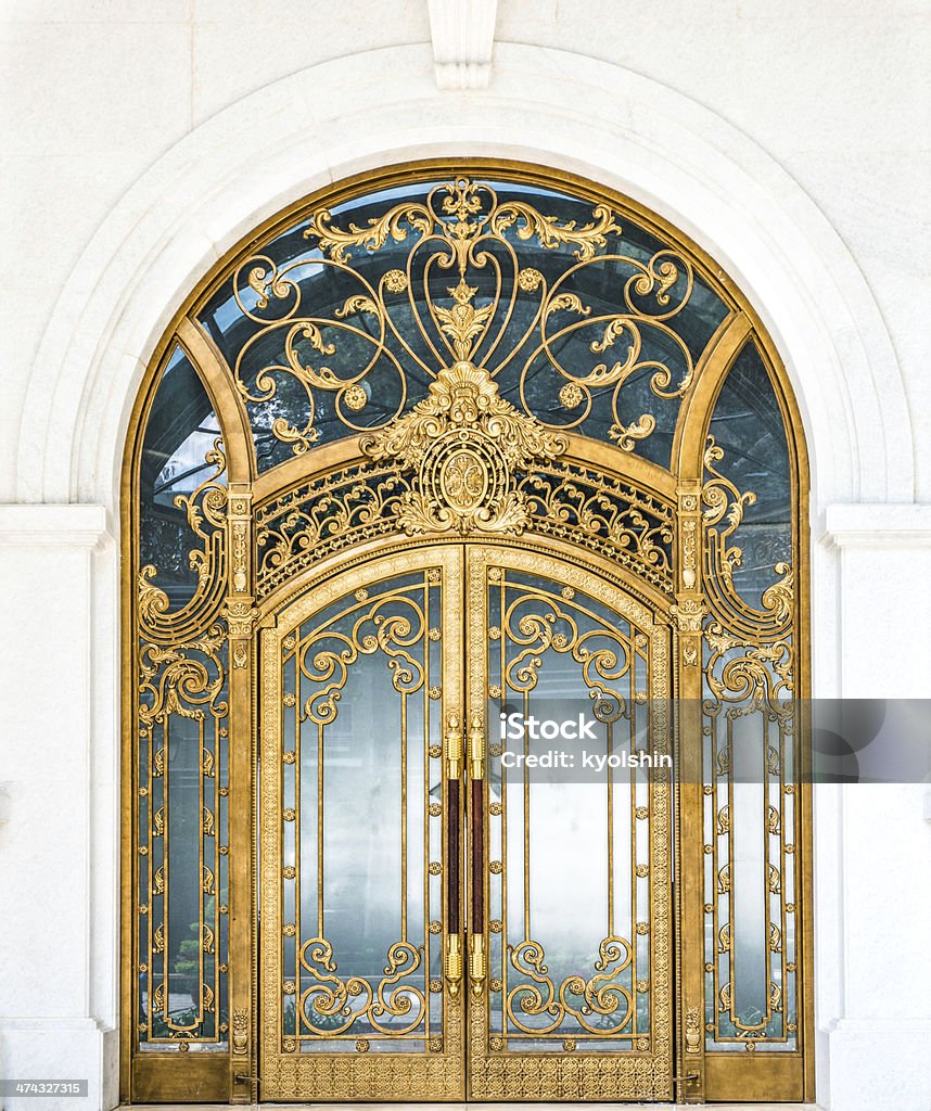 Closed door of building with gold ornate pattern. Beautiful arched doorway. Door made of wood, gold and glass reflecting arch. White wall of building with elegant gilded entrance. Old style of door with golden ornate details. Tourist attraction. Gold Colored Stock Photo