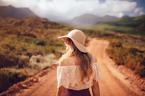 Cropped rearview shot of a boho girl in a cream coloured felt hat standing on a deserted dirt road on a summer day