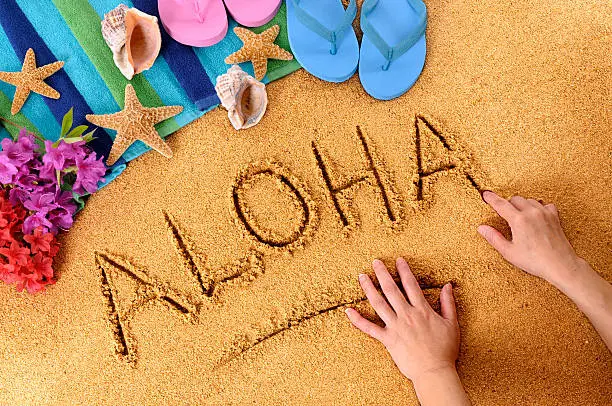 Child writing the word Aloha on a sandy beach, with flowers, beach towel, starfish and flip flops.