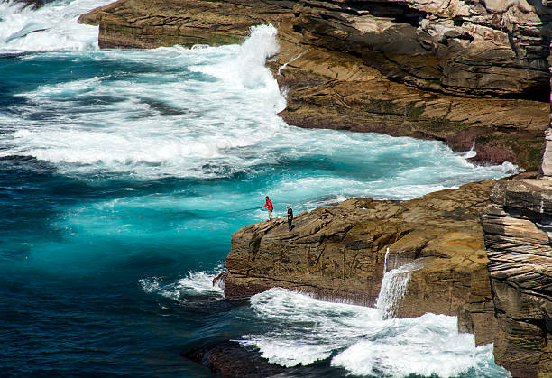 Sydney, Australia – April, 06 2015: Fishermans balances on a cliff many meters above sea level at time of rock fishing in Diamond bay, Sydney, Australia