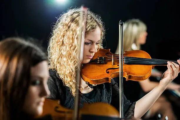 Three women playing cello, piano and violin and reharsing.  Focus is on serious young woman playing violin.