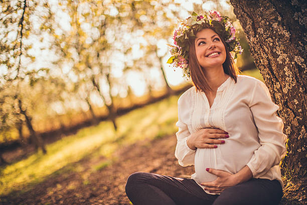 Photo of a pregnant woman relaxing in nature and sitting under a cherry blossom tree, wearing spring flowers head wreath