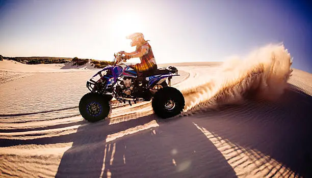 Competitive quad biker kicking up a plume of sand while racing over a sand dune