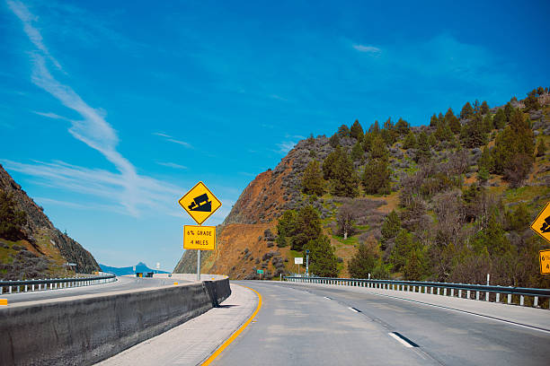 Warning road sign ahead of the upcoming downhill for semi truck mounted on a multi highway with a border separating the hills covered with trees, drifting into the horizon.
