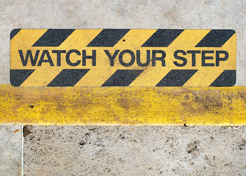Warning sign on concrete steps down to the river bank of the Thames in Surrey, England: WATCH YOUR STEP. The background is highlighted in yellow, with broad black diagonal lines.