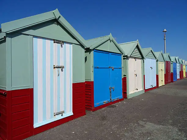 Photo showing a row of wooden beach hut sheds in Brighton and Hove, located in the county of East Sussex, England, UK.  These beach huts have a traditional seaside character, being painted pale green, with brightly coloured doors.